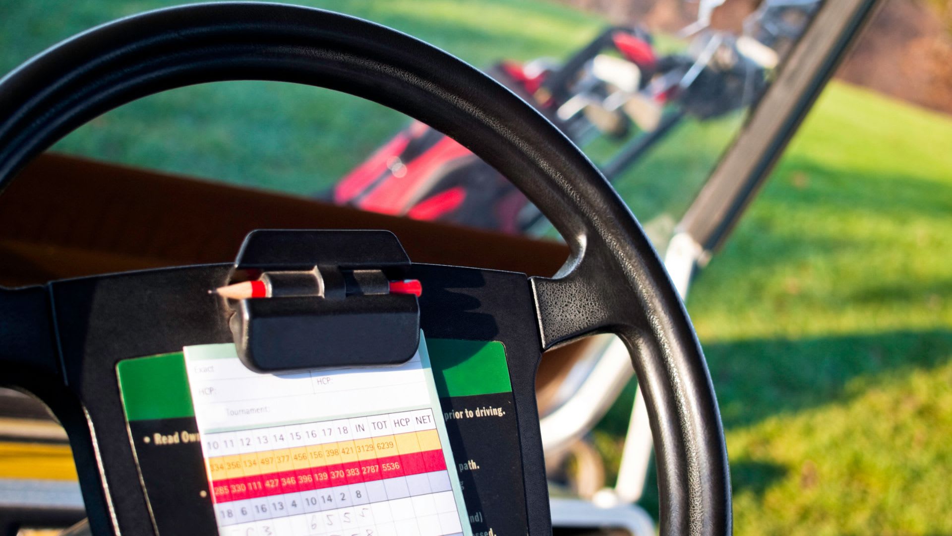 A golf cart steering wheel with a pencil and scorecard on a clipboard highlights golf scoring; a golf bag and green grass are visible in the background.
